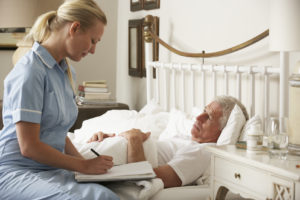 Nurse Visiting Senior Male Patient In Bed At Home Writing Notes.